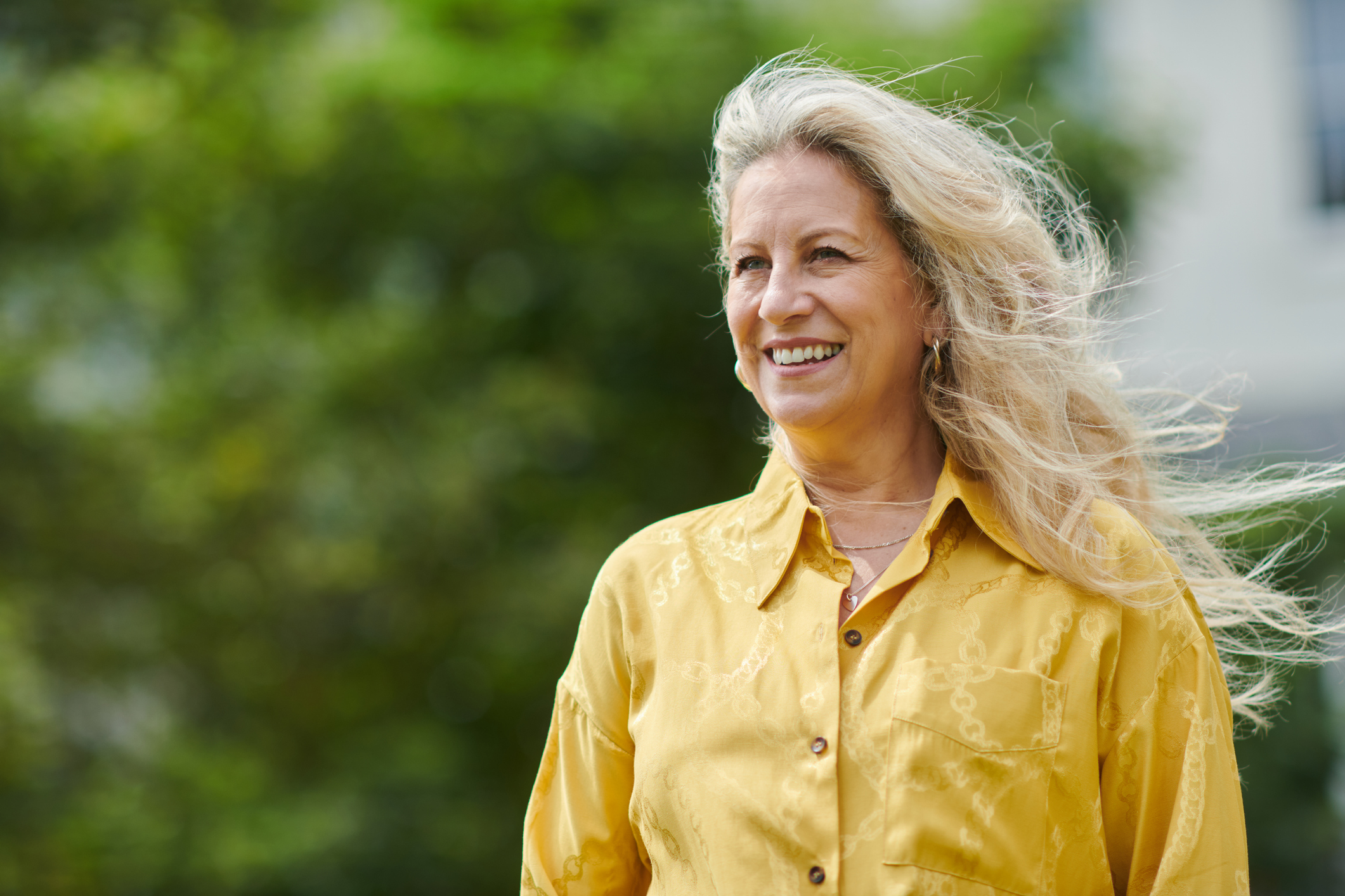 Vrouw met geel shirt en mooi blond haar kijkt lachend uit over haar tuin