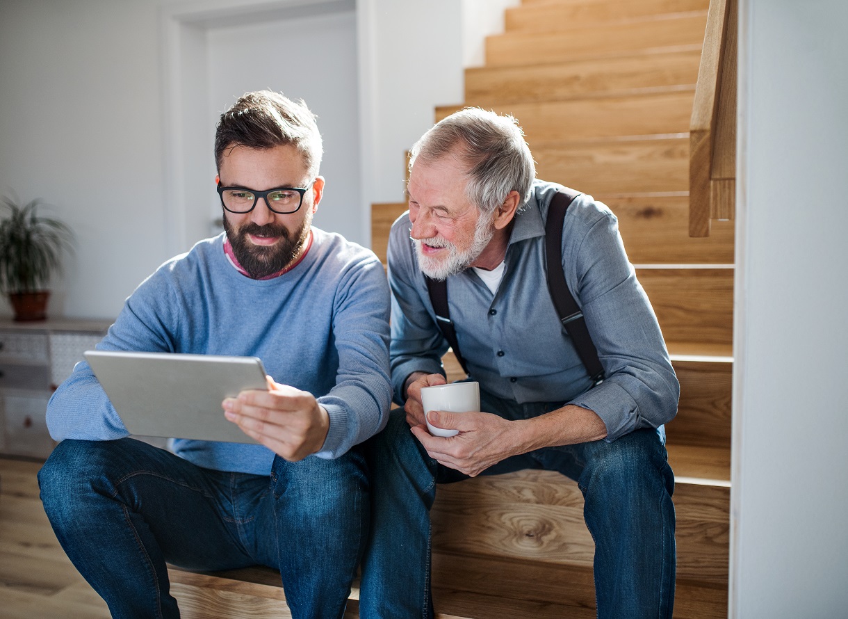 An adult son and senior father with tablet sitting on stairs indoors at home.