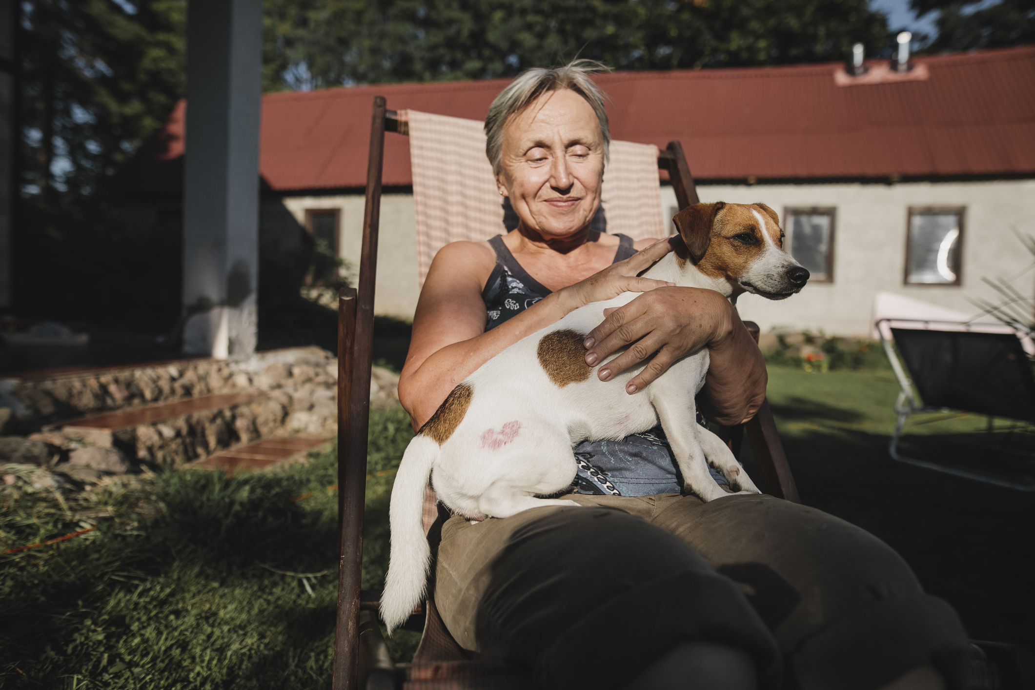 Vrouw zit tevreden op tuinstoel in de zon met hondje op schoot, huis op de achtergrond zichtbaar