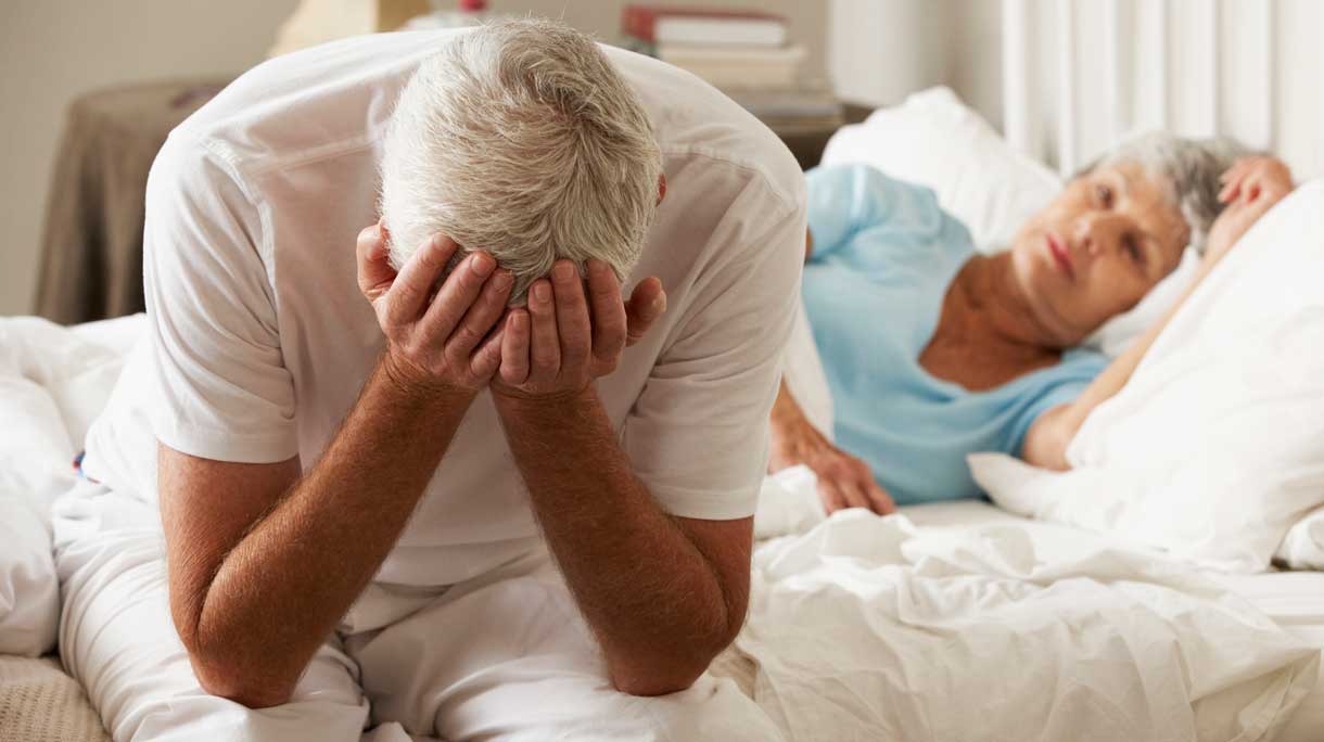 Man met handen voor zijn gezicht in bed met vrouw op de achtergrond