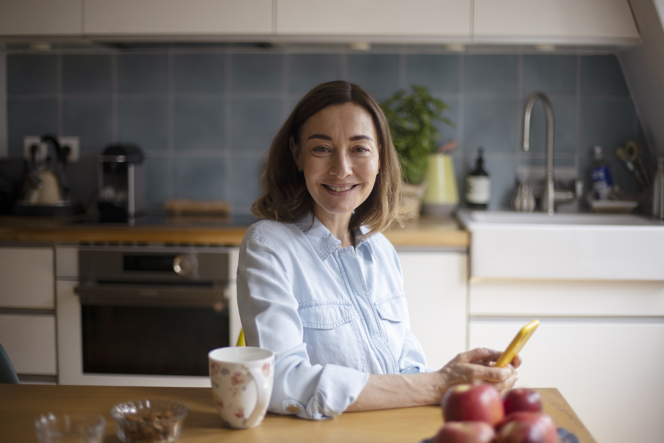 Vrouw zit in de keuken met smartphone, kijkt in camera